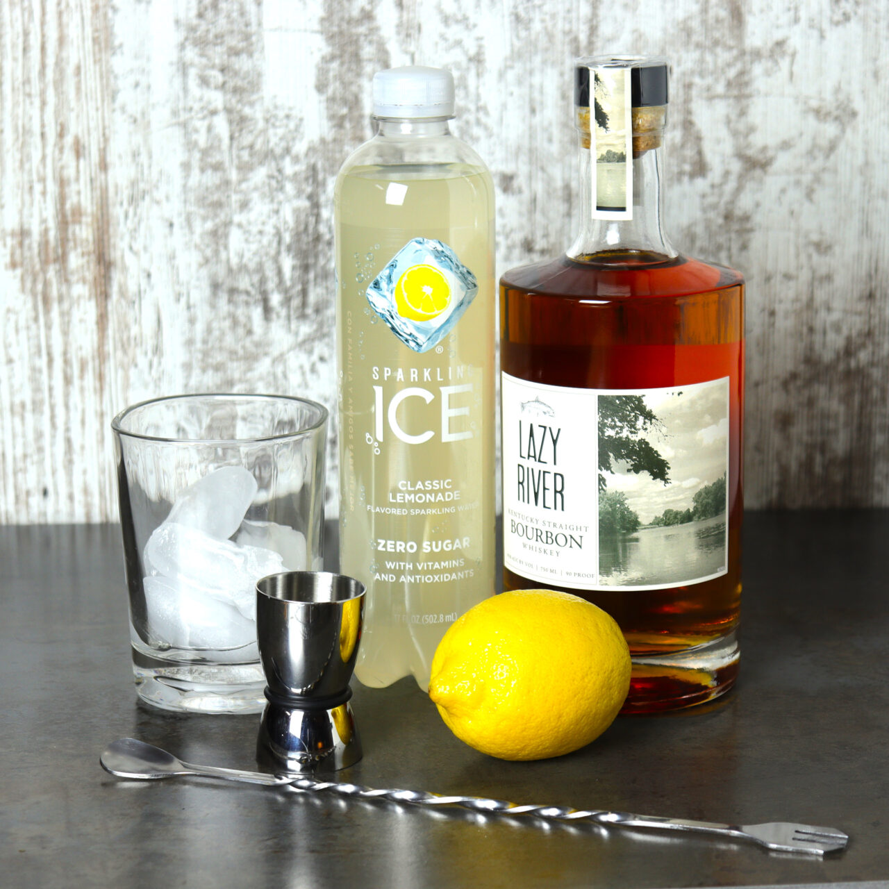 Ingredients for a whiskey lemonade cocktail, including a rocks glass, ice, lemonade, bourbon and lemon.