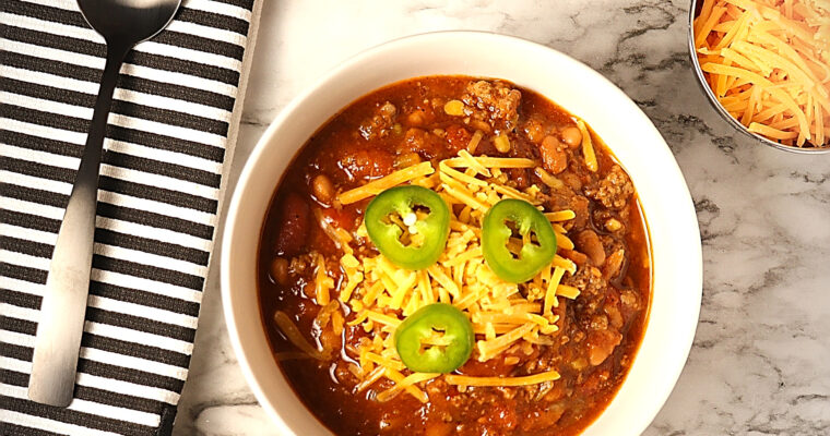 Rich warm bowl of chili, generously topped with melted cheddar cheese and adorned with fresh slices of jalapeño.