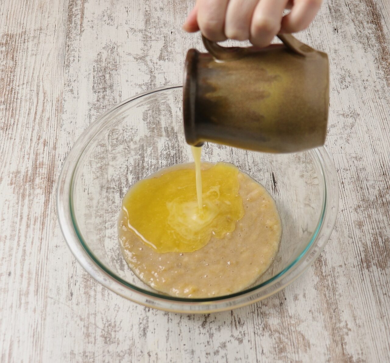 Pouring melted butter into a mixing bowl of mashed bananas.