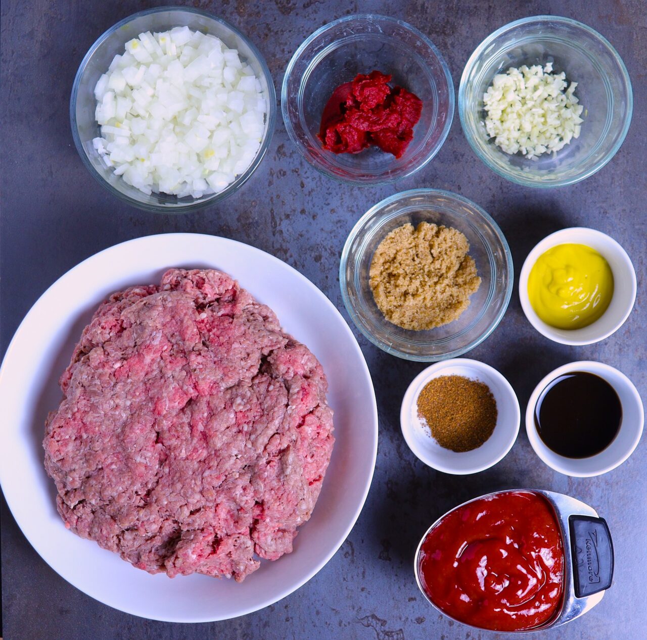 A vibrant display of fresh and colorful ingredients for scratch-made sloppy joes. Includes ground beef, diced onion, minced garlic, tomato paste, ketchup, yellow mustard, brown sugar, chili powder, and Worcestershire sauce.