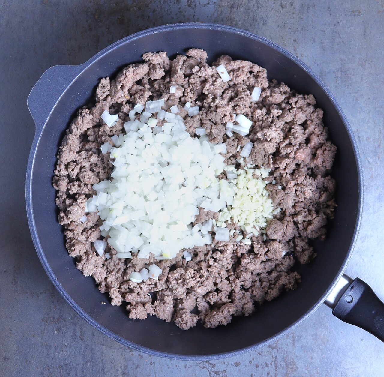 A close-up image of browned ground beef in a skillet, topped with freshly added diced onions and minced garlic. The onions and garlic are just starting to soften, adding a pop of freshness and aroma to the sizzling beef.