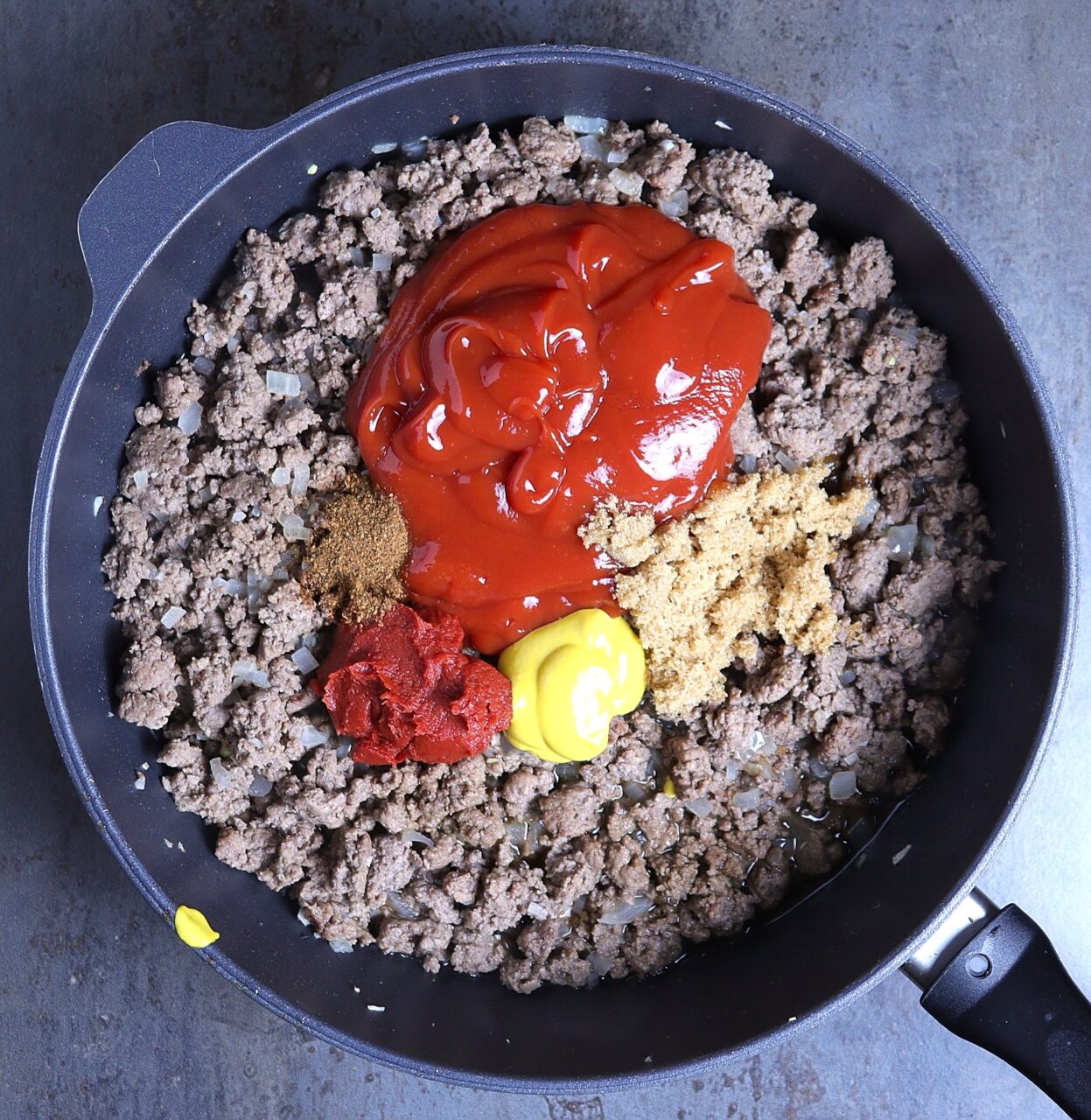 A skillet filled with browned ground beef, diced onions, and minced garlic, topped with vibrant layers of tomato paste, ketchup, brown sugar, Worcestershire sauce, yellow mustard, chili powder, and water. The ingredients sit in distinct sections, ready to be stirred together into a rich, flavorful sauce.