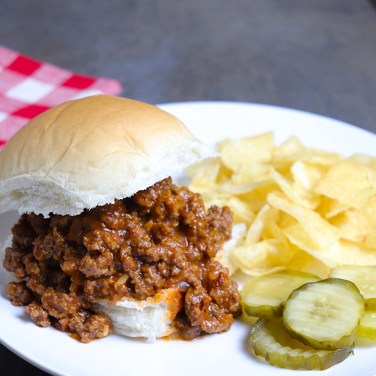 A mouth watering sloppy Joe sandwich filled with rich, savory meat sauce, serve on a soft bun alongside crispy potato chips and a few tangy pickle chips on a white plate.