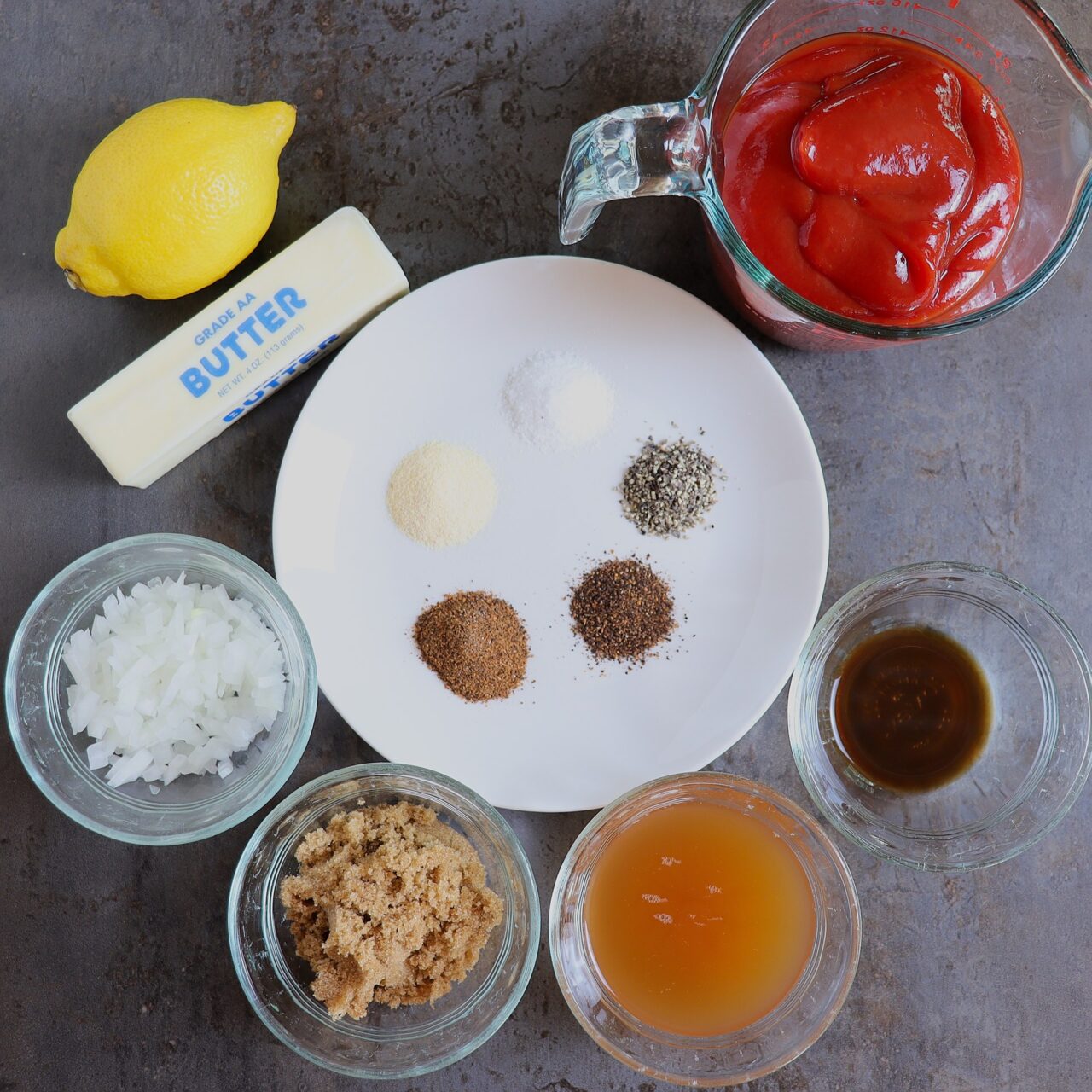 Ingredients: butter, diced onion, ketchup, apple cider vinegar, brown sugar, garlic powder, chili powder, Ancho chili powder, Worcestershire sauce, lemon, kosher salt, and black pepper, arranged neatly on a countertop.